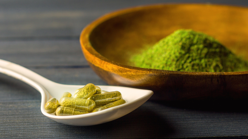 Loose green kratom powder in a wooden bowl with capsules of kratom capsule dosage in the foreground.