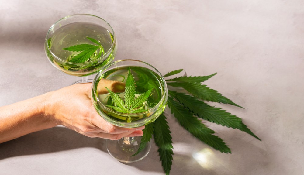 CBD with alcohol? It’s safe. This woman’s hand is holding beverage glasses with hemp leaves inside.